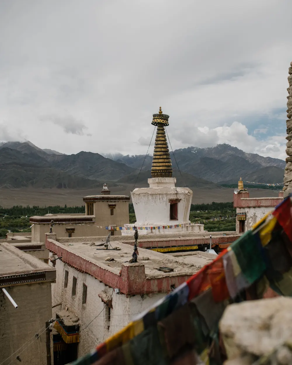 Roof of ancient temple with shabby walls with tower and hanging flags against cloudy sky and mountains in distance in town