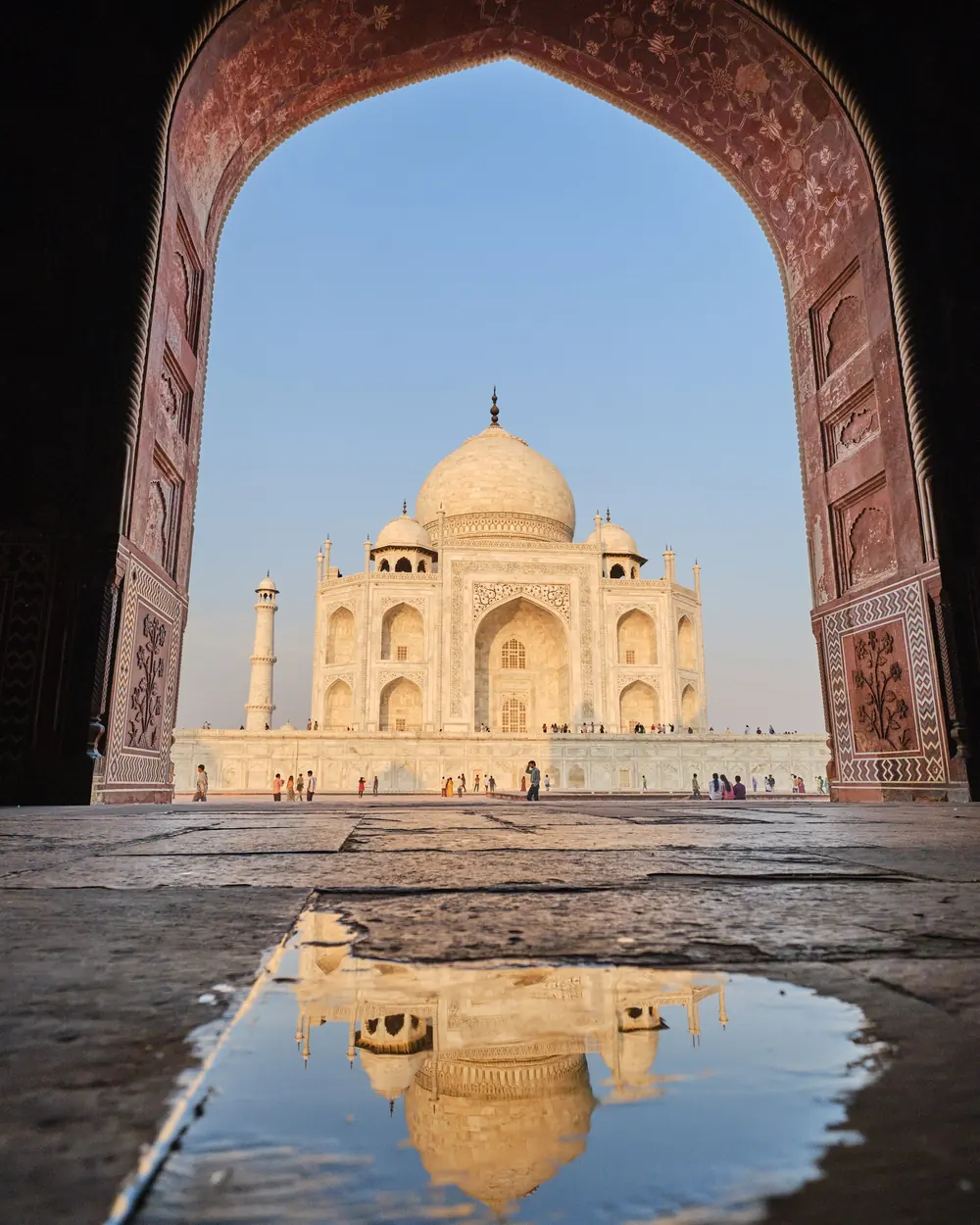 A stunning view of the Taj Mahal framed by an ornate arch with a reflection in a puddle